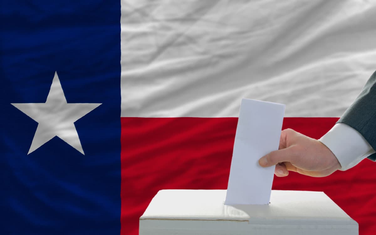 A hand casting a ballot into a box in front of the Texas flag.